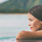 Luxury vacation woman relaxing in infinity swimming pool on summer travel at beach resort. Asian girl tourist on wellness spa relaxation outside in nature landscape banner panorama.